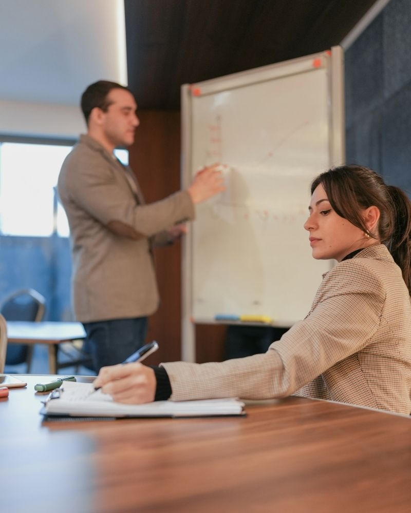 A woman sits at a table taking notes while a man stands at a whiteboard presenting or explaining something in a modern office or classroom setting.