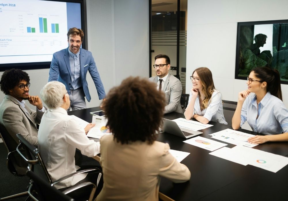 A group of six professionals sit around a conference table with laptops and documents, engaged in a discussion. One person stands and speaks to the group. A presentation with charts is displayed on a screen in the background.