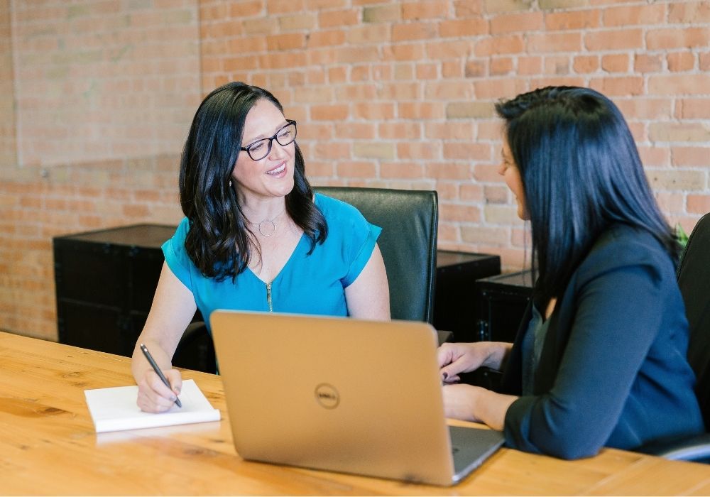 Two women sit at a wooden table in an office with exposed brick walls, talking and smiling. One has a notepad and pen, while the other sits in front of an open laptop.
