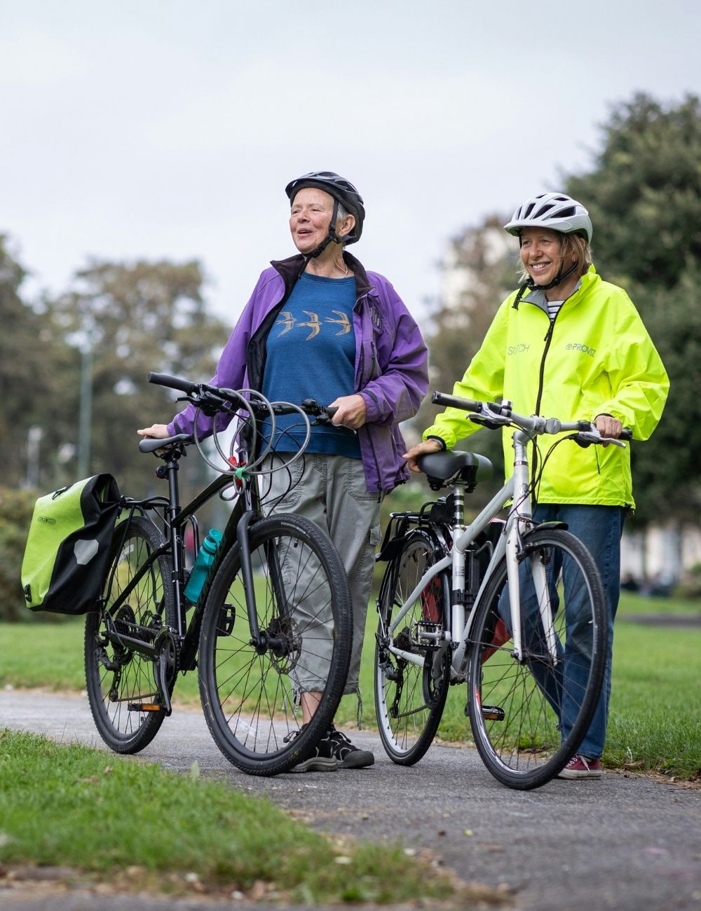 Two older adults wearing helmets and bright jackets stand outdoors with their bicycles on a paved path, smiling and looking to the side. Lush green grass and trees are visible in the background.