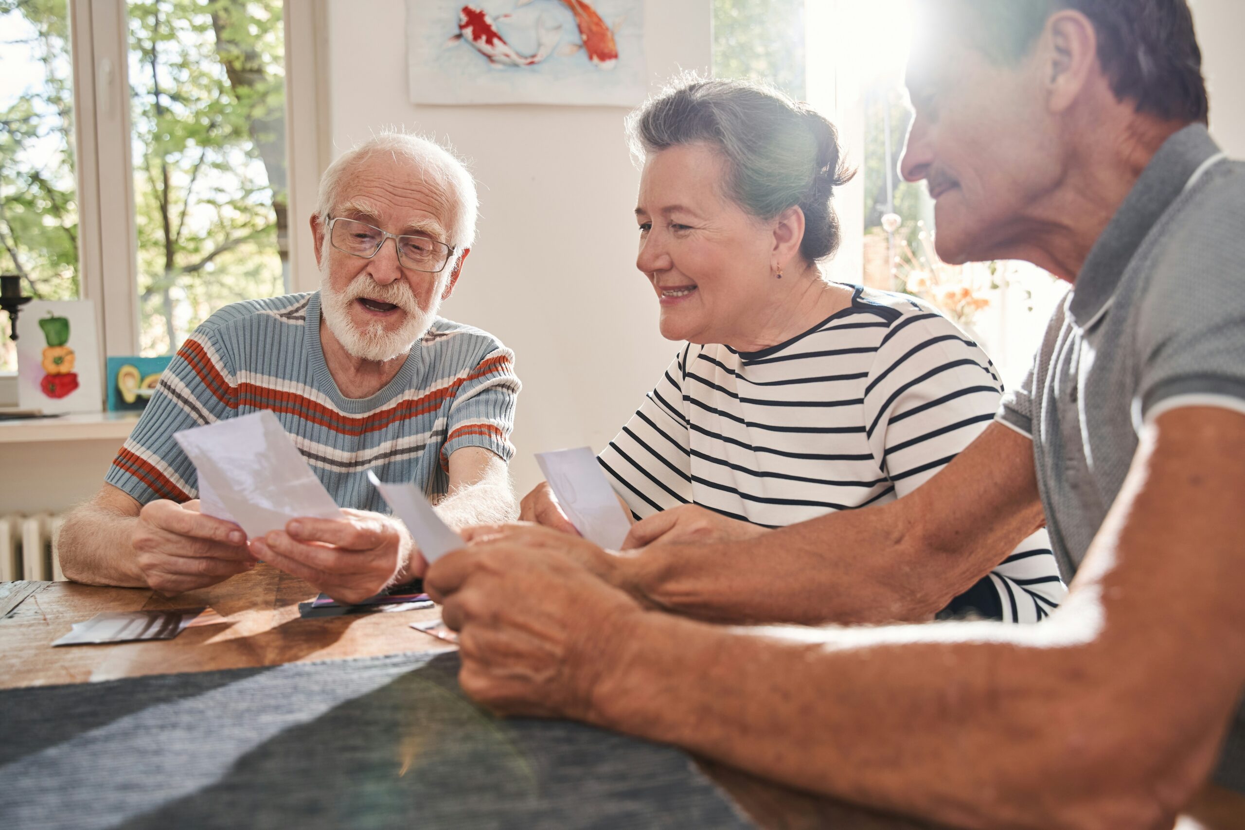 Three older adults sit at a table, smiling and looking at photographs together. Sunlight streams through the window behind them, creating a warm and cheerful atmosphere—an ideal moment to discuss long-term care planning in Arkansas.